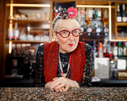 An older woman with red-rimmed glasses and a bow on her head pouts with her hands folded in front of her.