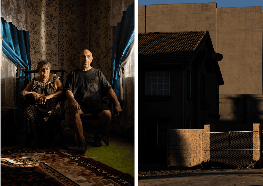 A man and a woman sit in the living room of their home. A large concrete wall erect behind a smaller home.