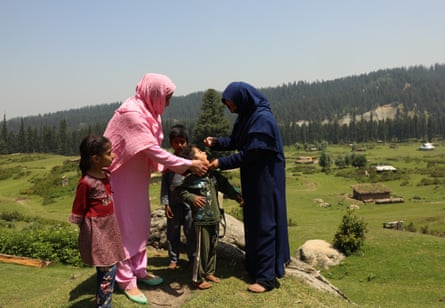 One woman holds a child by the chin as another administers drops into their mouth in a green rural setting. Two other children stand next to them