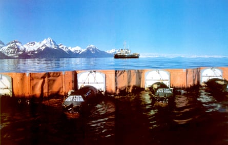 Orange and white floating line separates black oil from blue water, with snow-capped mountains beyond.
