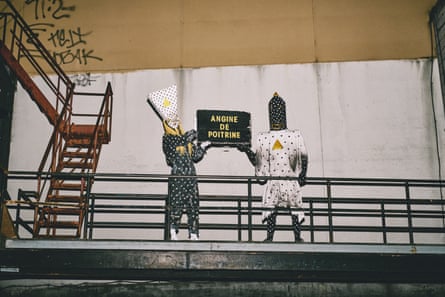 Two people dresked in polka-dot costumes wearing polka-dot headwear on a stage with a ladder in the corner. They are holding a sign saying ‘Angine de Poitrine’