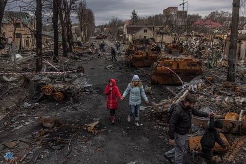 Two adults and two children in quilted jackets walk past the remains of military vehicles