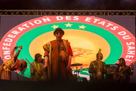 An African man in a long robe singing on stage with a guitarist behind him