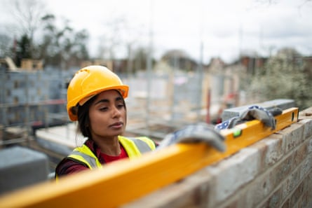 A young Asian woman in an orange hard hat uses a levelling tool on a brick wall.