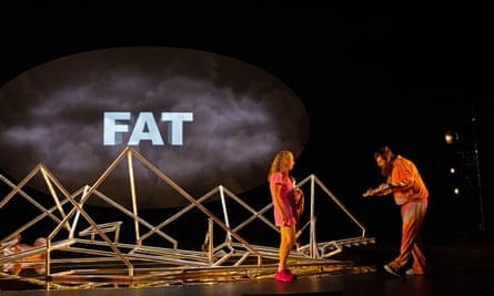 dark stage with actors next to large metal object and word “fat” projected on wall