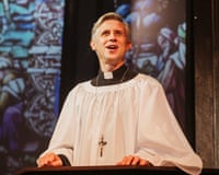 Actor Ian Hallard, in clergy robes, stands at a lectern with stained glass projected behind him