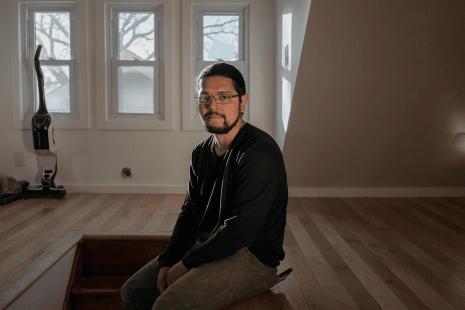 Aramis Rosa, a slender dark-haired, bespectacled man in light gray jeans and a long-sleeved black t-shirt, sits for a portrait in his renovated attic, with three narrow windows behind him.