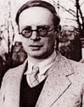 Grainy contemporary portrait of Voigt, clean shaven with slicked down hair and heavy-rimmed round glasses, in a shirt and tie with a v-necked sweater and jacket