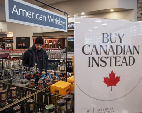 A man wearing a coat and beanie hat peruses bottles beneath a shop sign that reads 'American whiskeys', next to a poster that reads 'Buy Canadian instead'