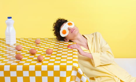 A woman leaning on the kitchen table with fried eggs over her eyes. There are whole eggs on the table, which has a gingham yellow cloth on it