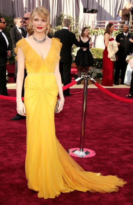 Michelle Williams in a bright yellow floor-length gown with a deep V-neckline stands on a red carpet at a formal awards event, holding a small clutch and wearing a silver statement necklace.