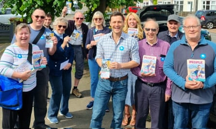 a group of mainly middle-aged and older men and women stand around their local MP, a younger man in his early 40s dressed casually in a checked shirt and jeans, in a leafy street holding up campaign material; they are all smiling in the sunshine.