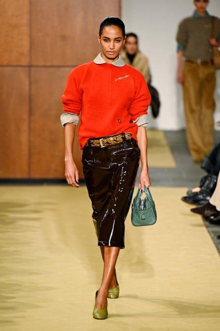 Model walks the Tory Burch runway wearing an orange jumper, patent leather skirt, and green heels