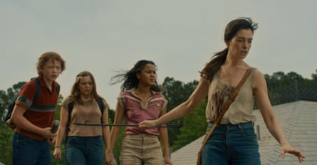 Anne Hathaway, holding a metal rod, leads three teenagers walking outdoors near houses