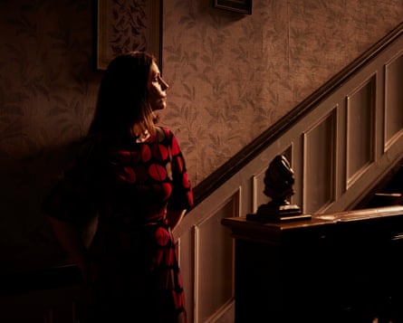 A woman stands half in shadow at the bottom of stairs in a house looking worried