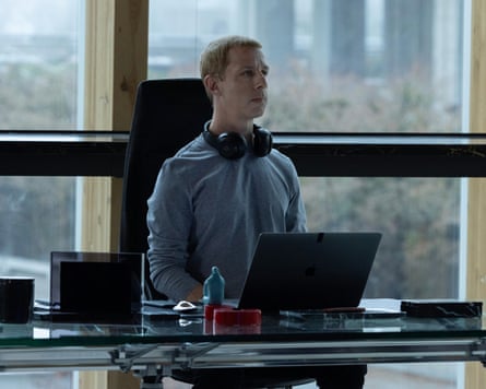 Jeremy Strong as Mark Zuckerberg, sitting upright at a laptop with headphones around neck and serious expression