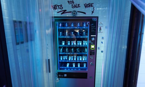 A photo of an NFT vending machine in Manhattan's Financial District is shown in glowing bluish and pinkish light. The machine looks like any you might see for drinks or candy, but above it is a scrawled sign reading "NFTS FOR SALE HERE."