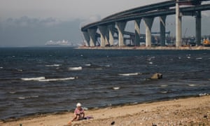 A women sits on a patch of beach and reads a book at Kanonerskiy Island, St Petersburg. In the background is the new bridge set to link the island with the mainland.