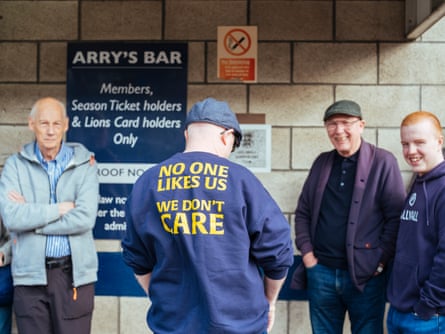A Millwall fan in a jumper that reads: ‘No one likes us we don’t care’ looks away from the camera as three other men look at the camera and laugh