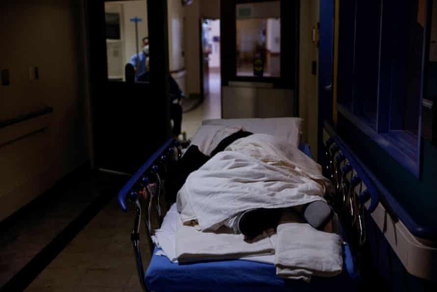 A patient lays on a medical gurney in a hallway waiting area of a hospital emergency department.
