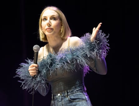 Katherine Ryan gesticulates on stage while wearing a fluffy top and holding a microphone.