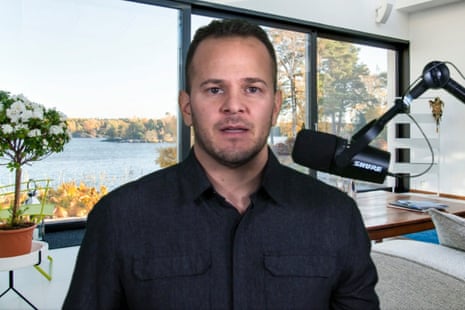 a man in a black shirt speaks into a microphone in a living room with a large glass window behind him
