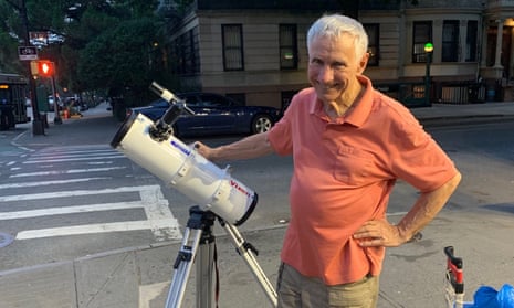An older white man wearing a salmon-colored polo shirt rests one hand on a telescope and the other on his hip, smiling at the camera