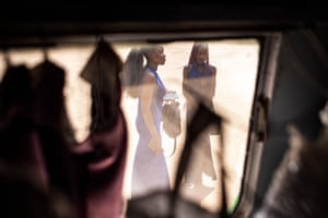 Two women in blue dresses stand holding their bags, seen through a window of the caravan