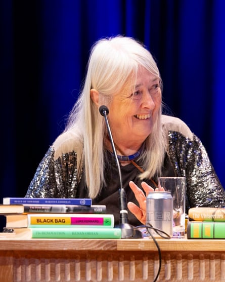Mary Beard sits at a wooden table behind a microphone and piles of books. She is in her early 70s with long, straight white hair. She is smiling and turning to look at a fellow judge.