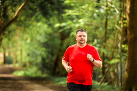 Color image depicting a man in his 30s jogging on a tree tunnel in a sunlit forest. The man is wearing sports training clothing - red t shirt, black shorts and trainers.