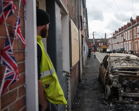 A gas utility worker looking out at burned cars on the street, from a house with UK flags pinned to it