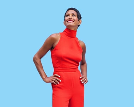 Naga Munchetty with her hands on her hips wearing a high-collar, sleeveless red jumpsuit, standing against a light-blue background