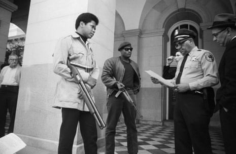 Two Panthers holding rifles speak to a police officer in a foyer.