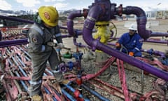 Two men in hard hats, jumpsuits and heavy boots work on dark purple painted pipes above, with pink floppy tubes and hard blue tubes.