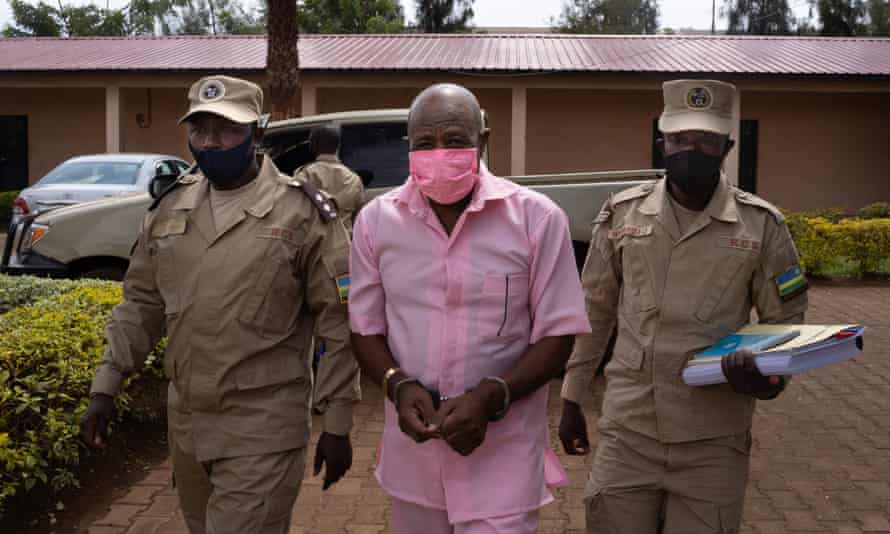 An older man, handcuffed and in pink prison clothing, is escorted by guards.