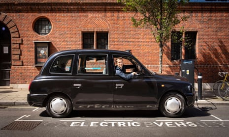 Black London cab plugged into to an electric charger with wall of red brick building behind.