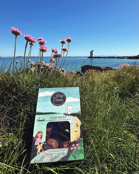 A small box of Islay Cocoa chocolates laid on grass next to flowers on a sunny day with blue sky and sea