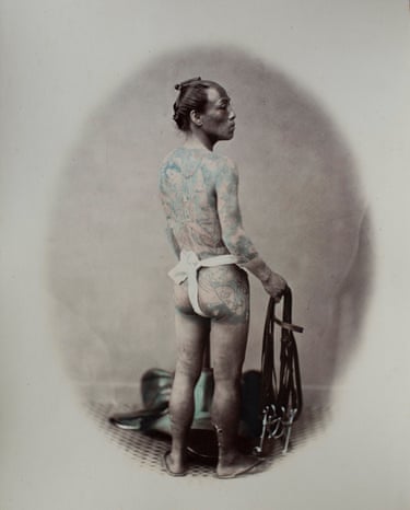 A heavily tattooed man stands bare-chested, looking back over his shoulder, holding leather straps