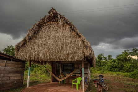 An open building with a thatched roof and a child lying in a hammock slung across the living area, under a sky darkened by rain clouds