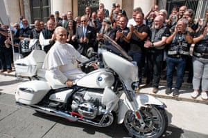 The pope sitting on a stationary, customised motorbike with bikers and other onlookers clapping