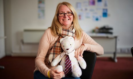 Portrait of Fay Turner in a jumper and scarf sitting smiling and holding a bear that is made out of white fabric with writing on it, and is wearing a miniature school tie