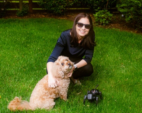 A woman in dark shades crouches down to stroke her dog. A black robot dog sits next to them on the grass