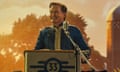 Kyle MacLachlan stands at a podium with a microphone against a hazy orange background