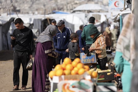 People shop at market stalls, with tents in the background and a display of oranges in the foreground