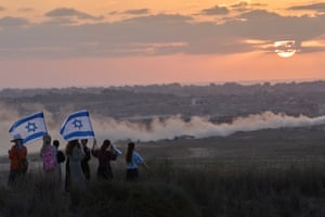 A group of people with Israeli flags look out from a hilltop towards Gaza with the sun low in the sky