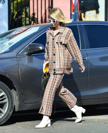 The actor Emma Roberts wearing a beige check co-ord, white ankle boots and sunglasses, walking past a car on a street.