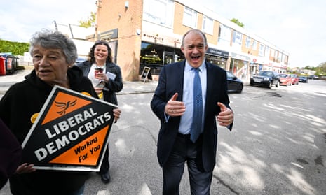 Ed Davey looking cheerful next to a party activist holding a Lib Dem placard as a woman holding a coffee looks on smiling behind them