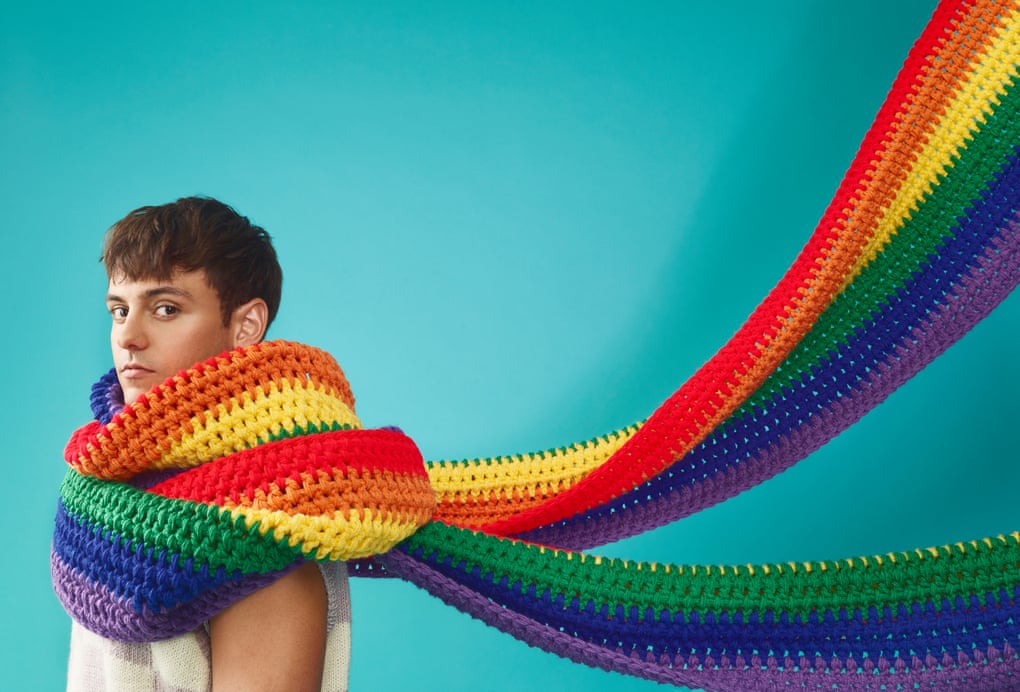 Tom Daley with an oversized crochet Pride scarf wrapped round his neck and streaming out behind him