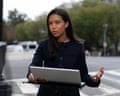 Woman in black outfit on street corner with laptop on table in front of her gestures as she speaks into a camera.