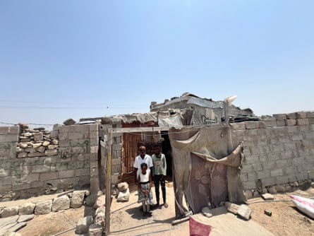 A older man and two children stand in a brick and fabric shelter